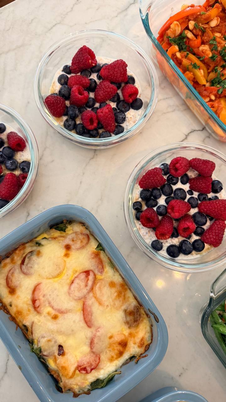 Glass bowls of chia yogurt overnight oats topped with fresh raspberries and blueberries, alongside a baked frittata with spinach and cherry tomatoes.