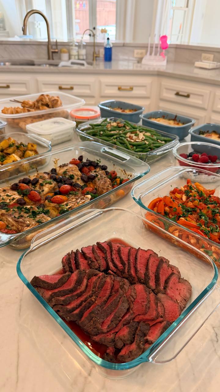 A kitchen counter filled with glass containers of sliced rosemary garlic filet mignon, Mediterranean baked cod with olives, almond green beans, and harissa peppers.