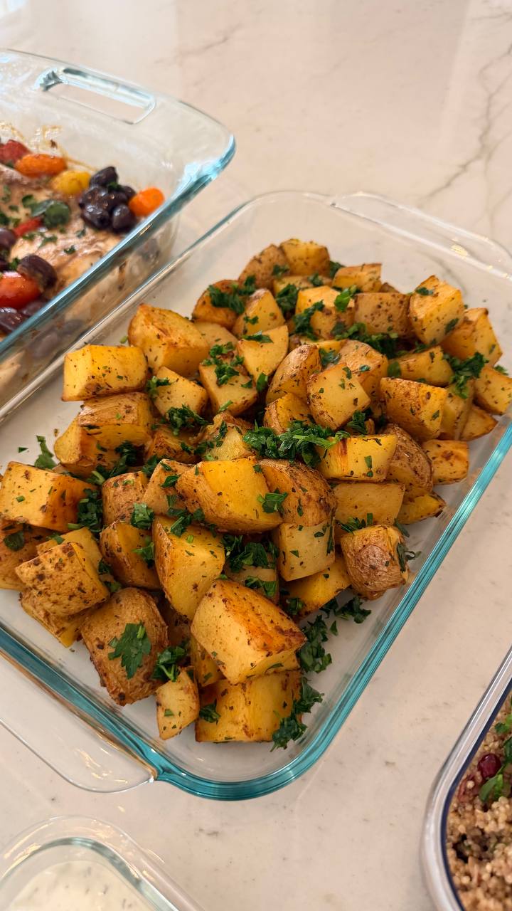 A glass baking dish filled with golden brown garlic and herb roasted potato wedges, garnished with fresh vibrant green chopped parsley.