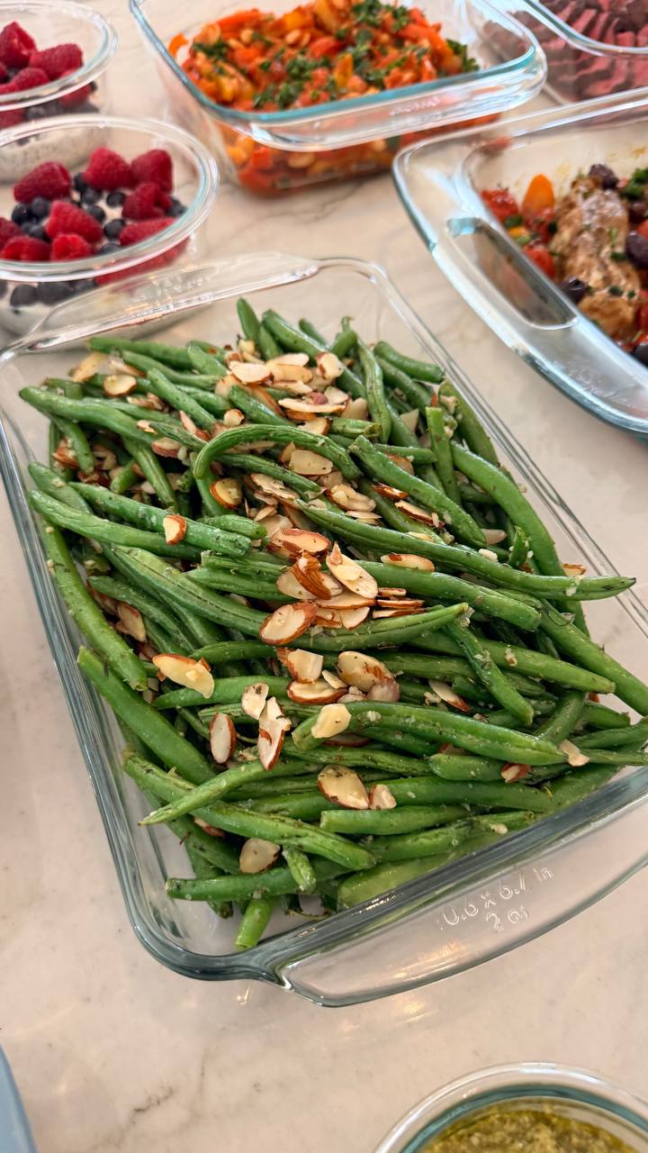 A glass serving dish filled with vibrant green beans almondine topped with toasted almond slices, presented alongside a spread of various meal prep containers.