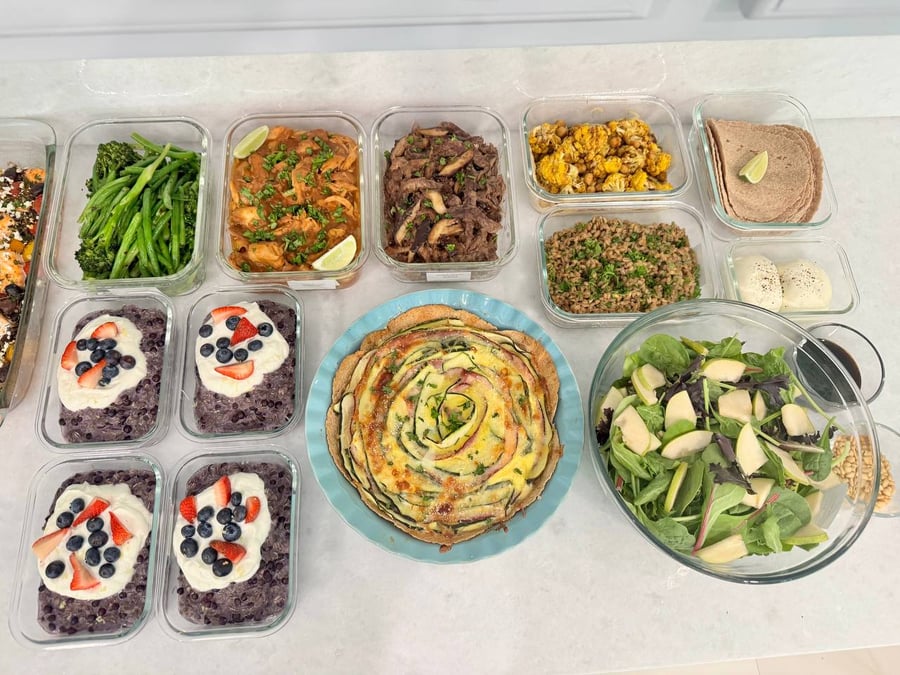 A variety of glass containers holding saucy chicken, beef sirloin, farro, blueberry oatmeal, a zucchini ham quiche, and fresh pear arugula salad.