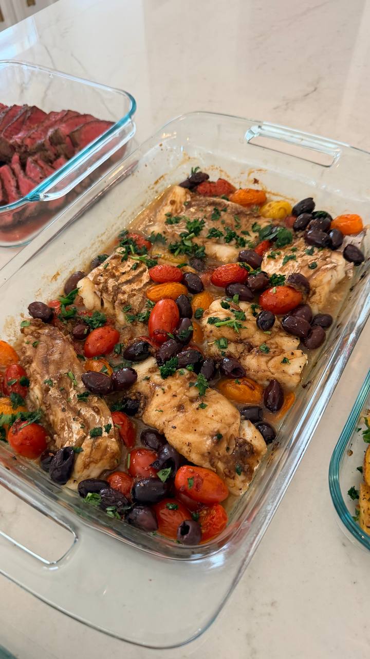 A glass baking dish containing mediterranean-style baked cod filets garnished with whole kalamata olives, cherry tomatoes, and a light sprinkling of chopped green herbs.