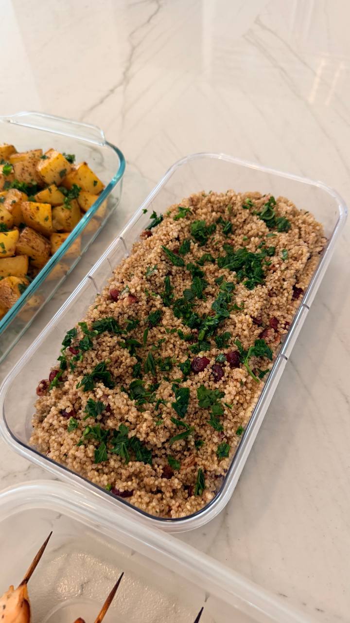 Glass containers featuring pecan and cranberry white quinoa garnished with fresh herbs alongside golden brown garlic and herb baked potato wedges on a marble surface.
