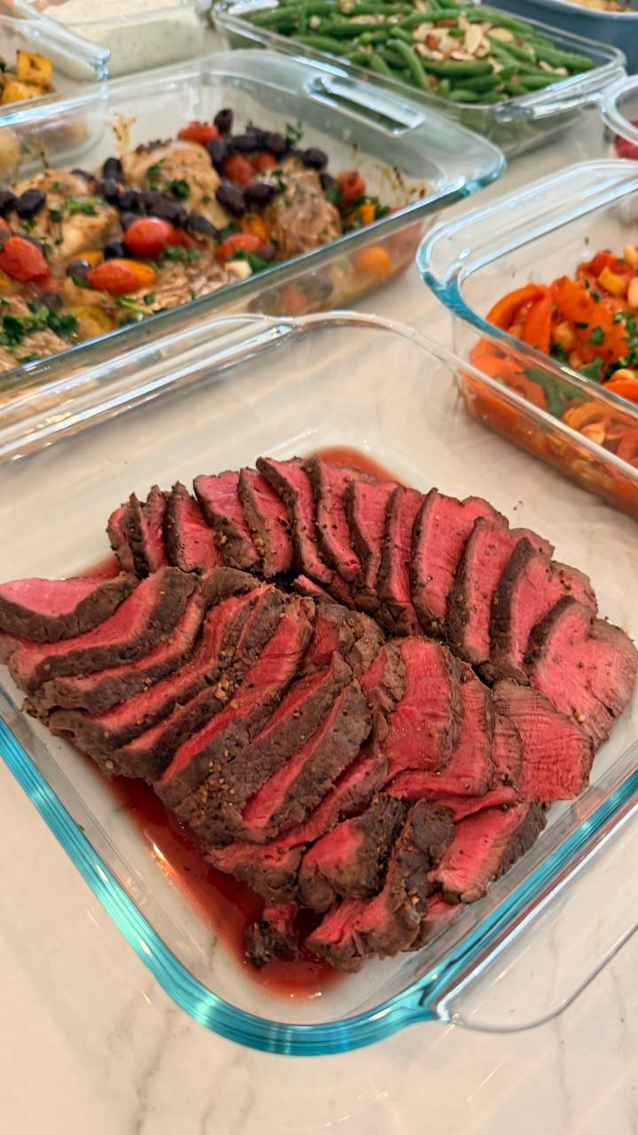 A glass dish containing thinly sliced medium-rare rosemary garlic filet mignon steaks, presented alongside other glass containers of Mediterranean cod, beans, and vegetables.