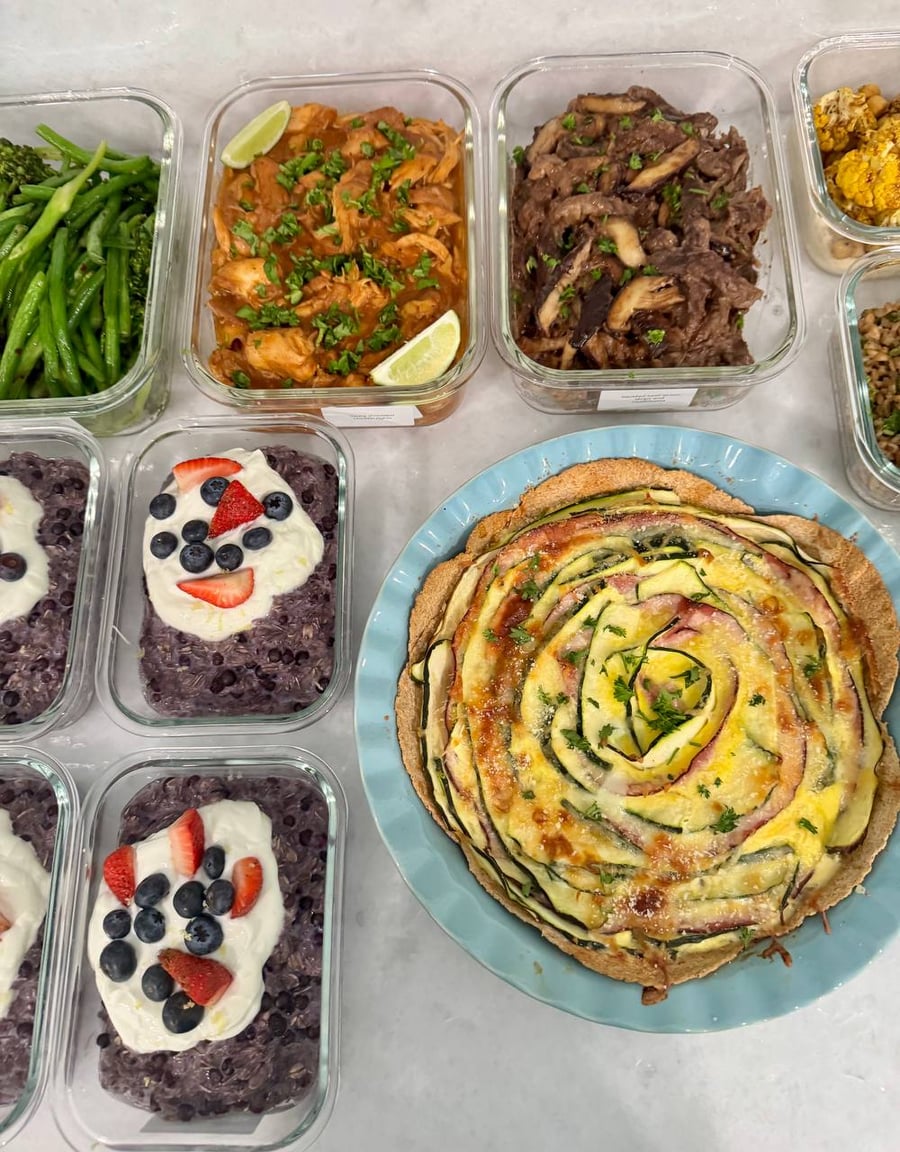 A freshly baked zucchini, ham, and tortilla egg quiche in a blue fluted dish, surrounded by various glass meal prep containers of nourishing food.