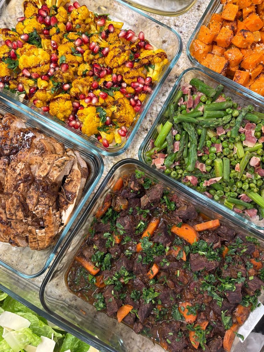 Glass containers featuring beef burgundy, mediterranean cauliflower with pomegranate seeds, balsamic chicken, garlic sweet potatoes, seared asparagus with pancetta, and fresh Caesar salad.