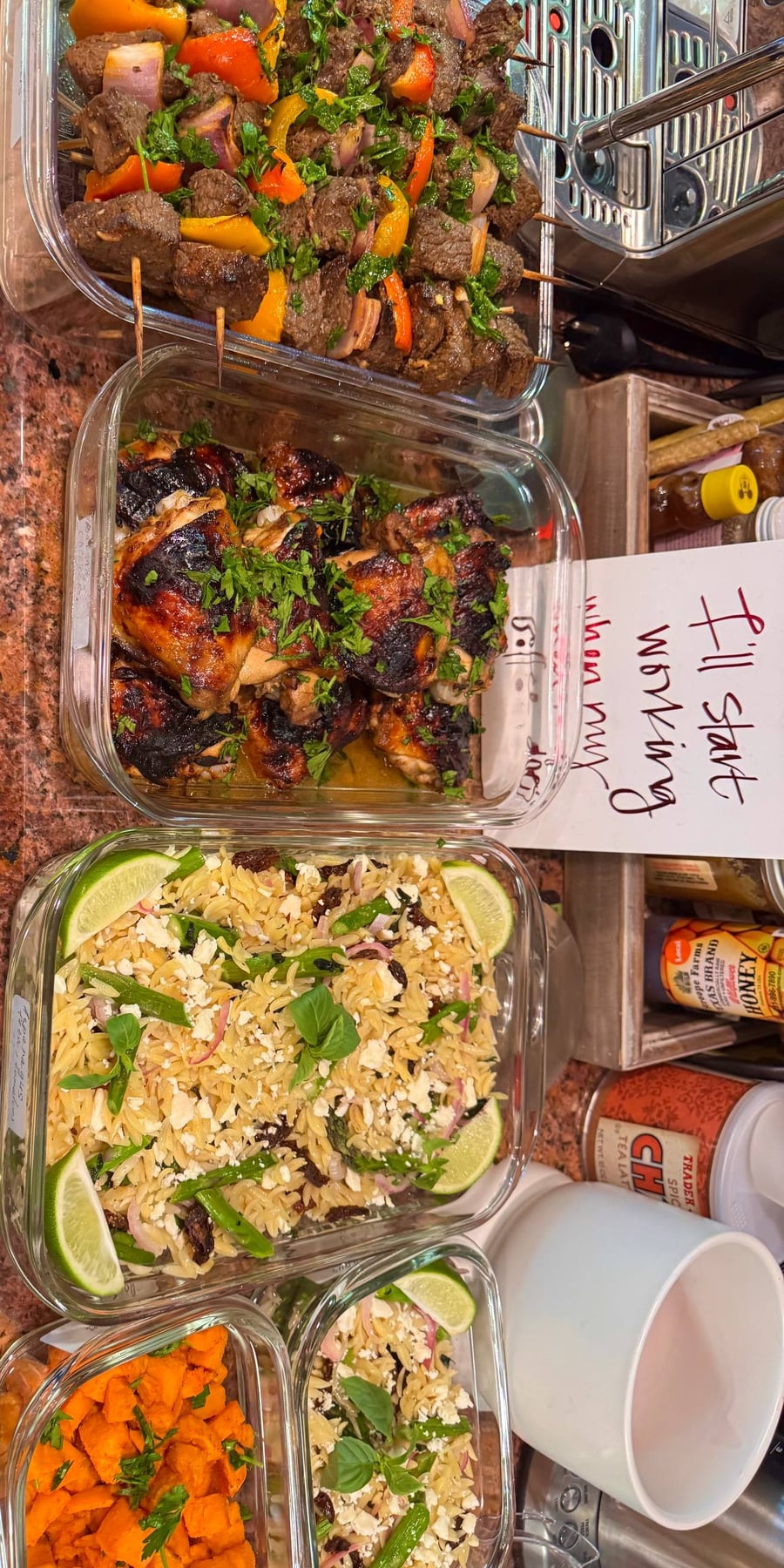 Glass containers holding beef skewers with peppers, balsamic glazed chicken thighs, and orzo salad with asparagus, sun-dried tomatoes, feta, and lime slices.