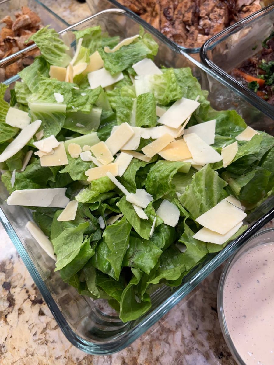 A glass container filled with fresh chopped romaine lettuce topped with generous shaved parmesan cheese, accompanied by a side of light creamy dressing.