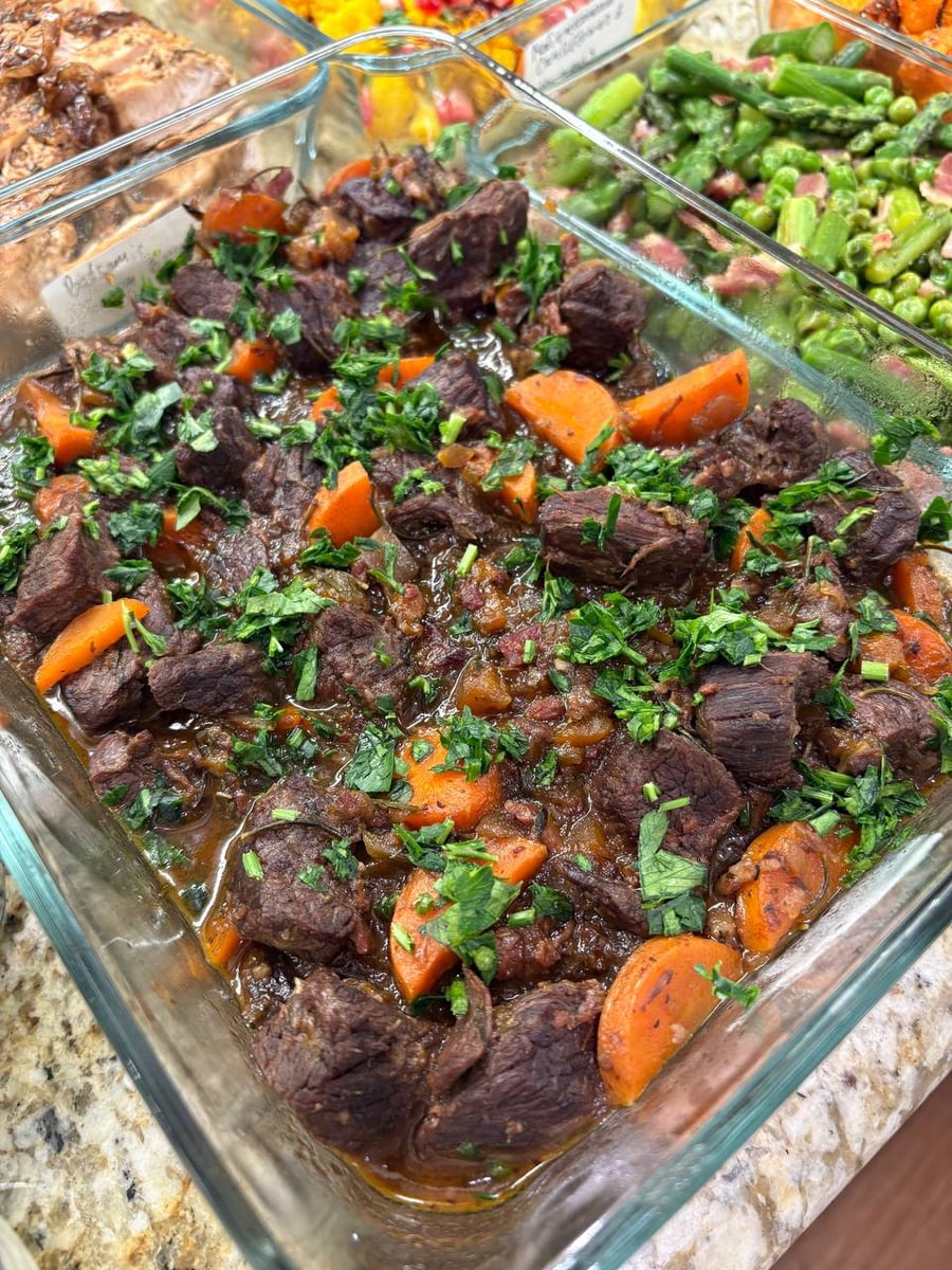 A glass container filled with hearty French beef Burgundy stew featuring tender beef chunks, bright orange carrot slices, and a dusting of fresh green herbs.