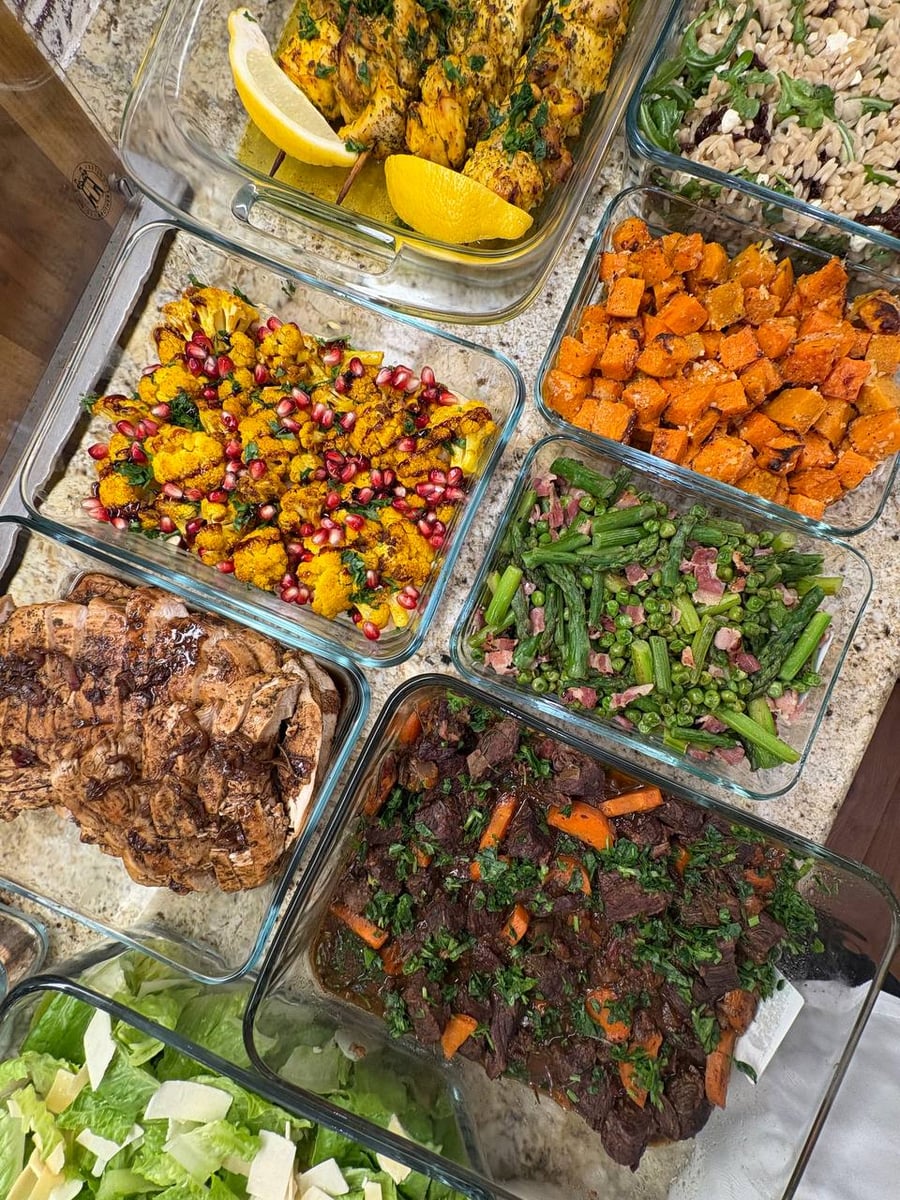 An assortment of glass containers filled with Greek chicken skewers, beef Burgundy, Mediterranean cauliflower, herbed sweet potatoes, asparagus with pancetta, orzo, and Caesar salad.