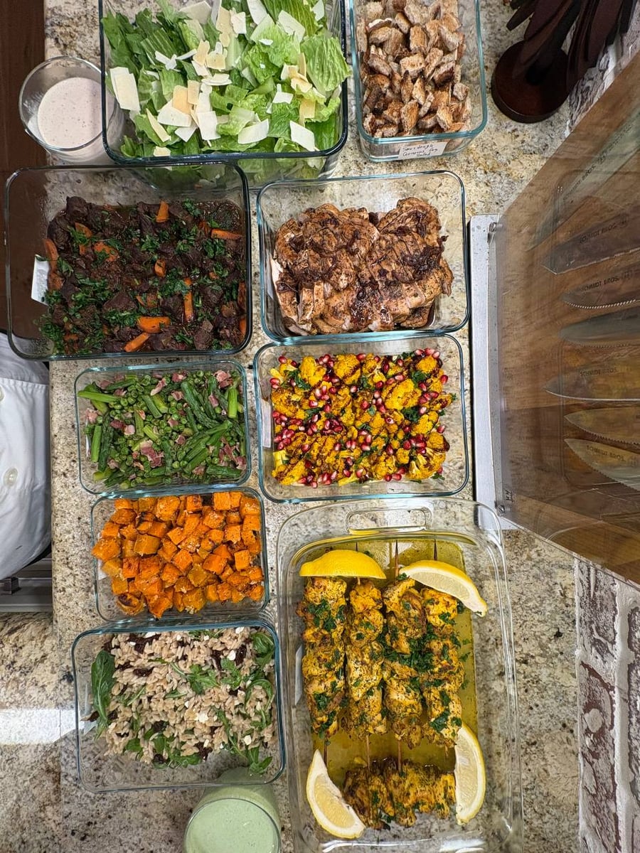 Top-down view of assorted glass meal prep containers featuring Greek chicken souvlaki, beef Burgundy stew, seared asparagus, roasted sweet potatoes, and various fresh salads.