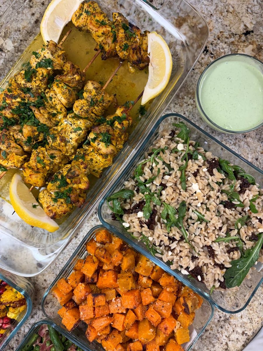 Glass containers holding Greek chicken souvlaki skewers, brown rice orzo with arugula and feta, and roasted sweet potato bites with a yogurt sauce.