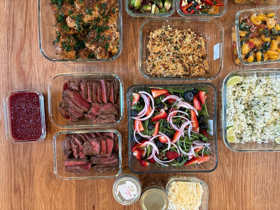 A variety of glass containers filled with balsamic chicken, pecan salmon, sliced filet mignon, gnocchi, berry arugula salad, and lime rice.