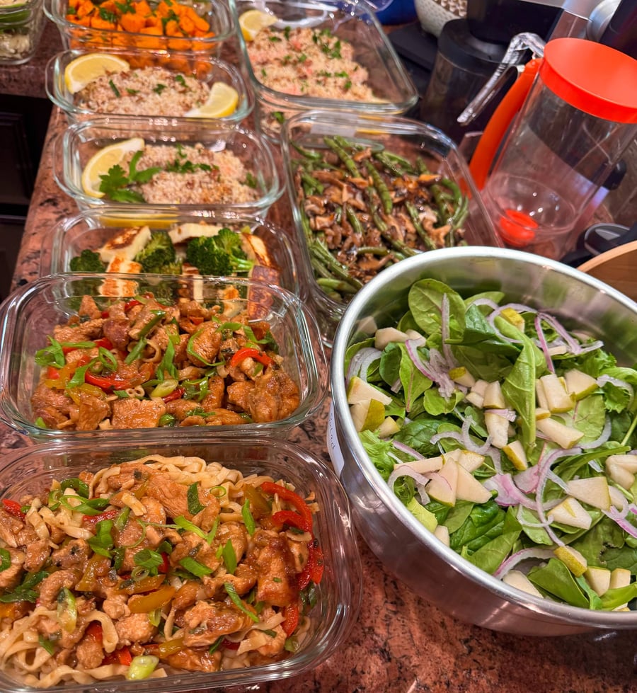A variety of glass meal prep containers filled with pecan-crusted salmon, chicken chow mein, sauteed broccoli with paneer, and a fresh pear spinach salad.