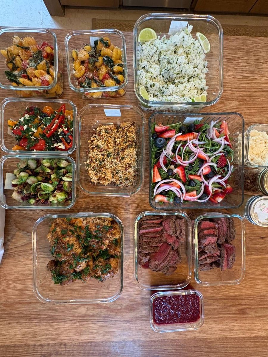 Glass containers holding pecan salmon, filet mignon, balsamic chicken, gnocchi, brussels sprouts, Mediterranean peppers, and a fresh berry salad on a table.