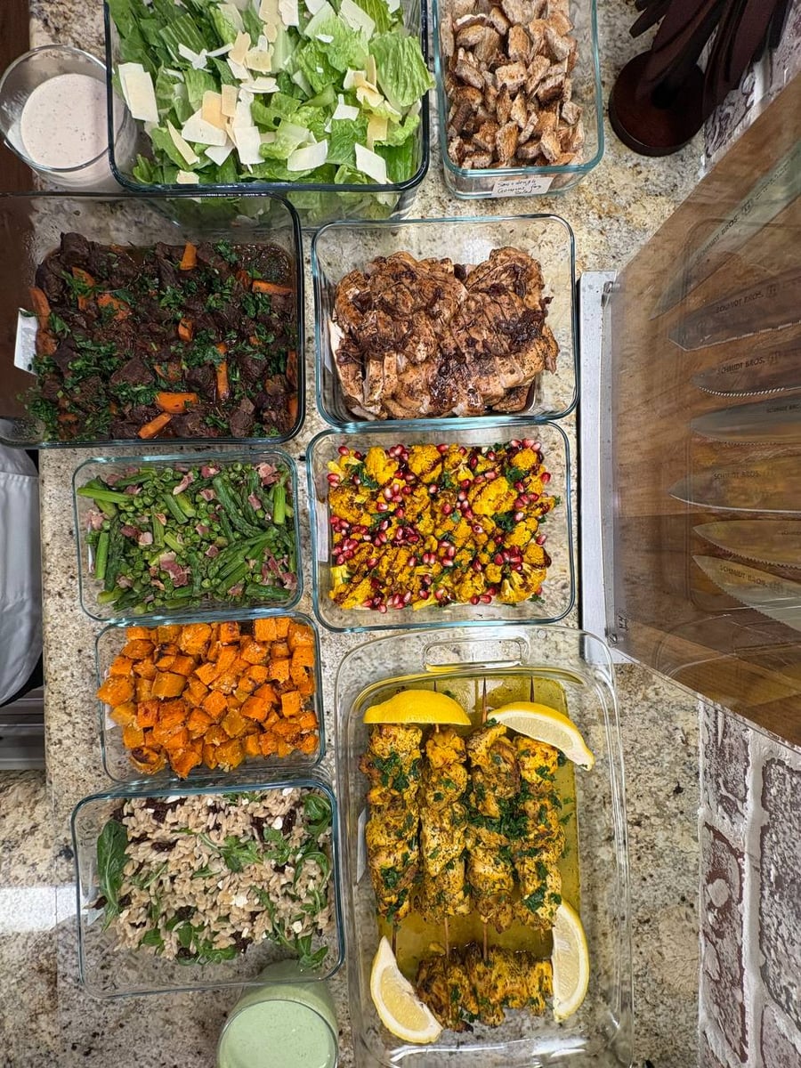 A variety of glass containers holding beef burgundy, balsamic chicken, mediterranean cauliflower, seared asparagus, orzo salad, caesar salad, and chicken souvlaki skewers.