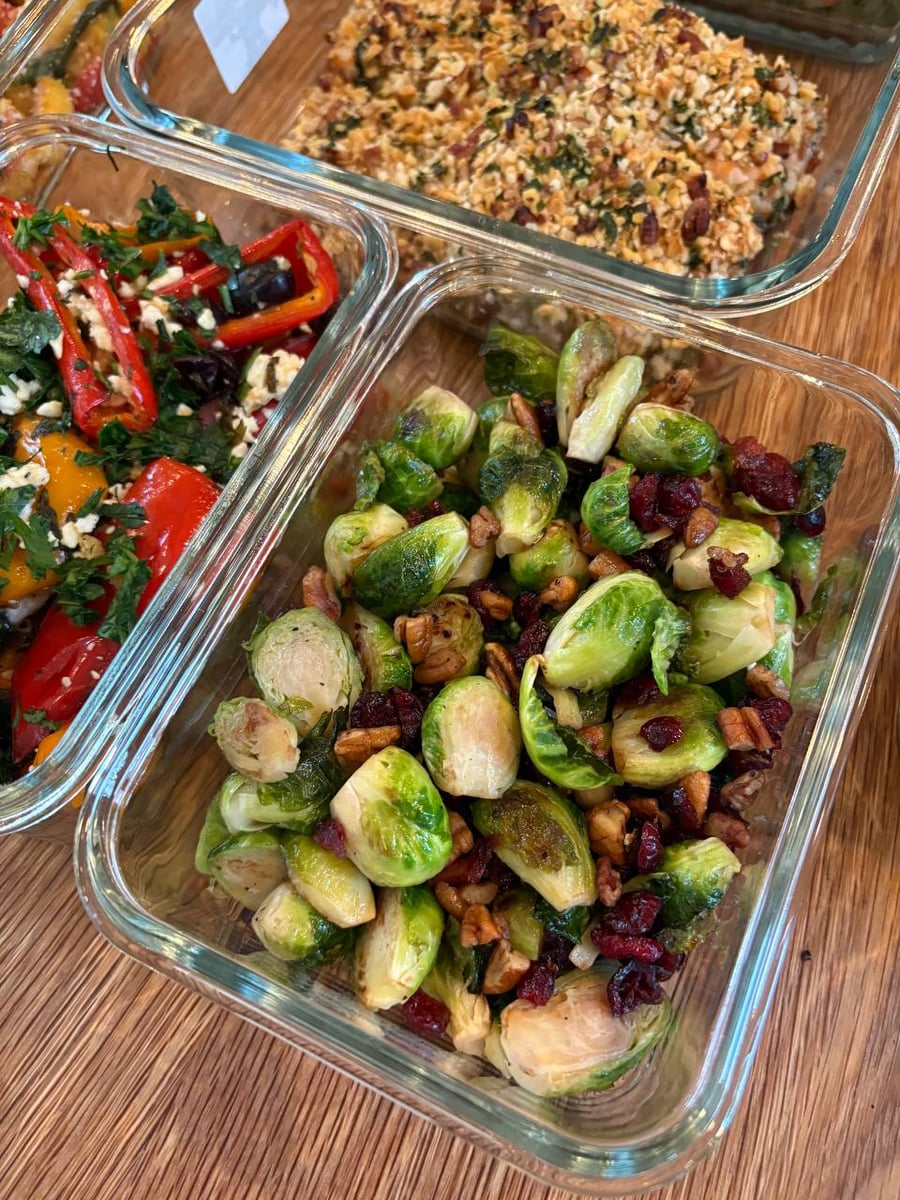 Glass meal prep containers filled with crispy pecan crusted salmon, mediterranean peppers with feta, and maple cranberry pecan seared brussel sprouts on wood.