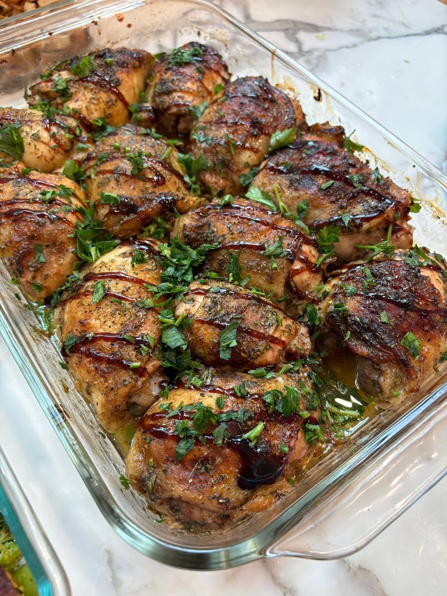 A glass baking dish containing braised balsamic chicken thighs, garnished with fresh chopped herbs and a dark, rich balsamic glaze drizzle.