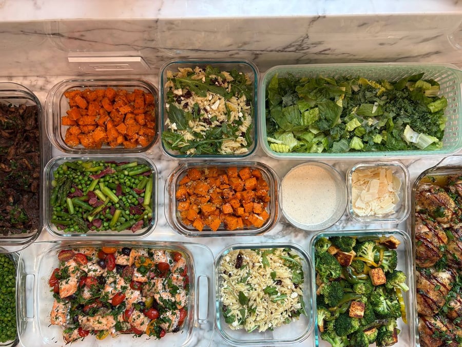 A variety of glass containers holding braised balsamic chicken, Mediterranean salmon, herbed sweet potatoes, seared asparagus, and fresh kale caesar salad with savory toppings.