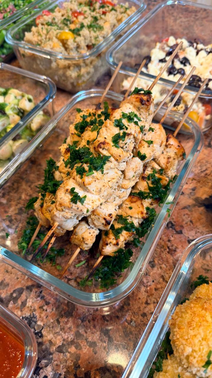 A glass container holds four Greek style chicken souvlaki skewers seasoned with herbs, presented alongside containers of quinoa salad and Mediterranean salmon.