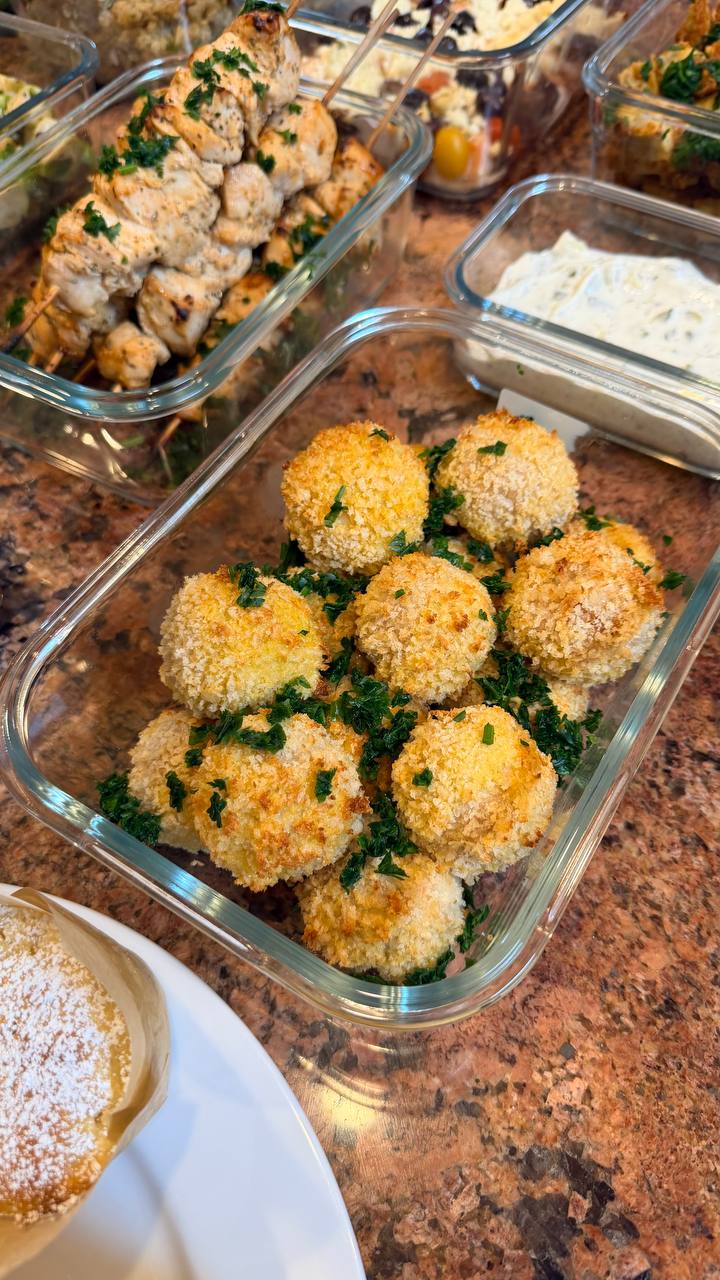 A glass meal prep container filled with crispy golden brown Kiev mozzarella meatballs garnished with fresh green herbs, displayed on a marble countertop.