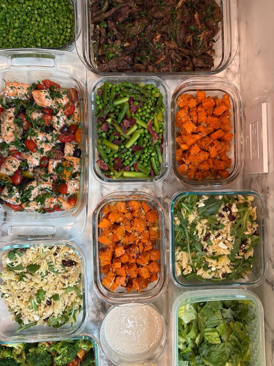 A variety of glass containers filled with Mediterranean salmon, beef sirloin with mushrooms, herbed sweet potatoes, and orzo salad served on a marble surface.