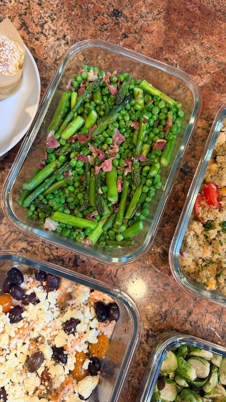 A glass container with seared asparagus, peas, and pancetta sits beside baked salmon with feta, olives, quinoa salad, and a raspberry white chocolate muffin.