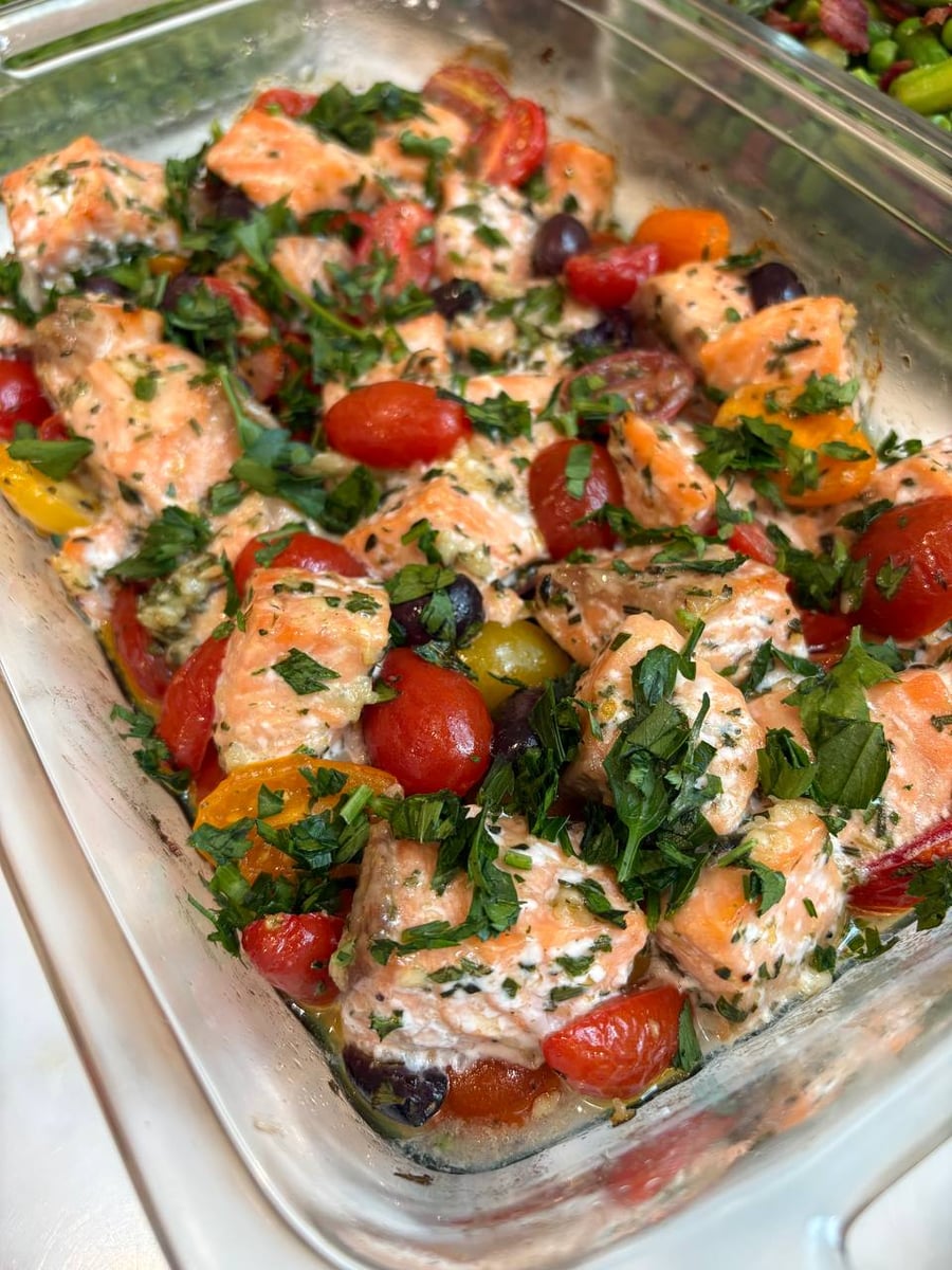 A glass container holds Mediterranean salmon bites, bright red and yellow cherry tomatoes, and dark Kalamata olives, all garnished with freshly chopped green parsley.