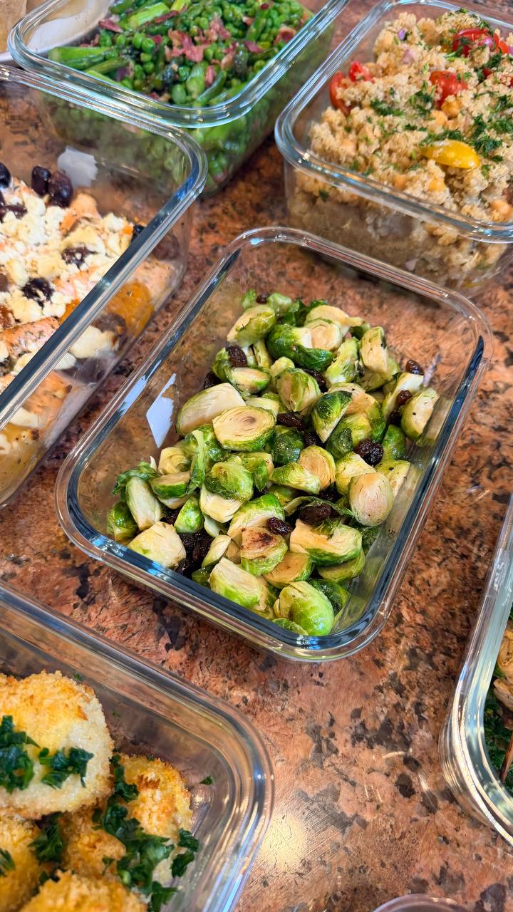 Glass meal prep containers filled with maple brussels sprouts, seared asparagus with pancetta, quinoa salad, mediterranean salmon bites, and breaded kiev mozzarella meatballs.