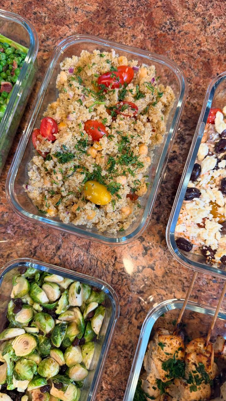Glass meal prep containers filled with quinoa chickpea salad, Mediterranean salmon bites with olives, and herb-grilled chicken skewers next to roasted Brussels sprouts.