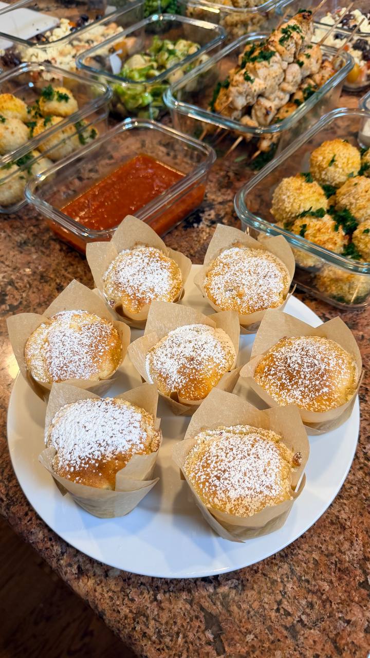 A white plate topped with raspberry white chocolate muffins next to glass containers filled with breaded mozzarella meatballs and chicken souvlaki skewers.