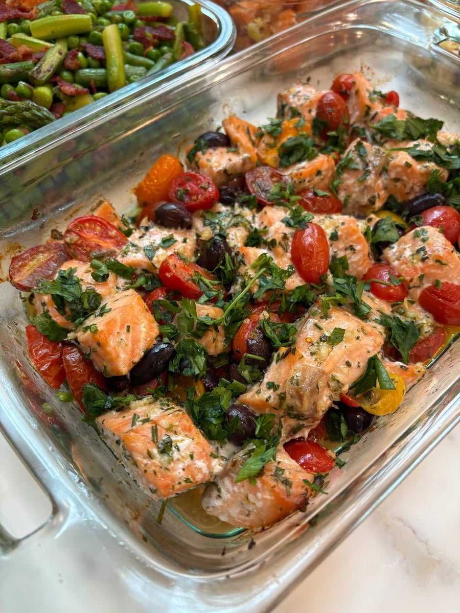 A glass tray filled with Mediterranean salmon bites, bright cherry tomatoes, and dark Kalamata olives, topped with fresh green herbs and served alongside vegetables.