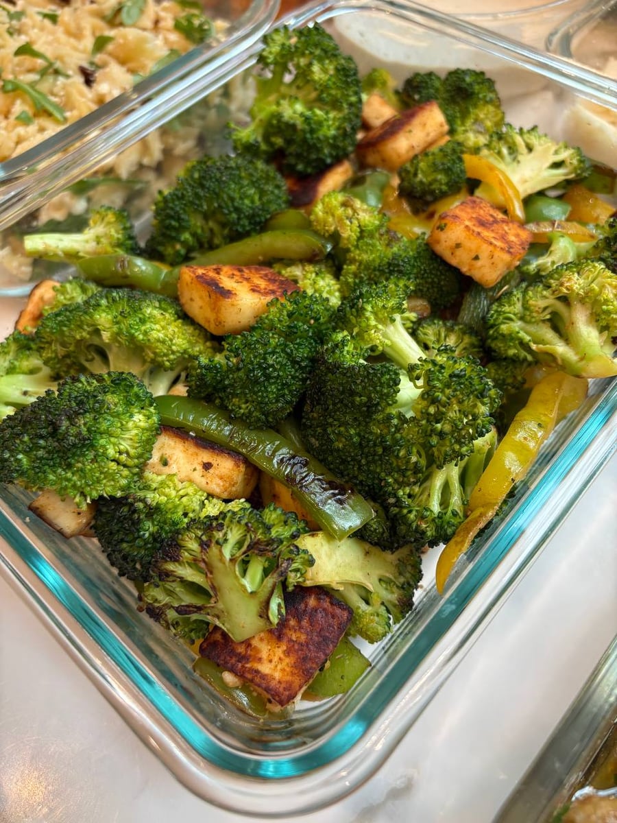 A glass meal prep container filled with sauteed broccoli florets, sliced yellow and green bell peppers, and cubes of golden-brown seared paneer cheese.