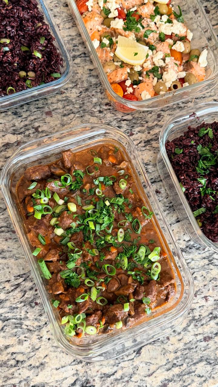 Glass meal prep containers filled with rich beef burgundy stew and separate portions of black rice topped with fresh scallions and herbs.