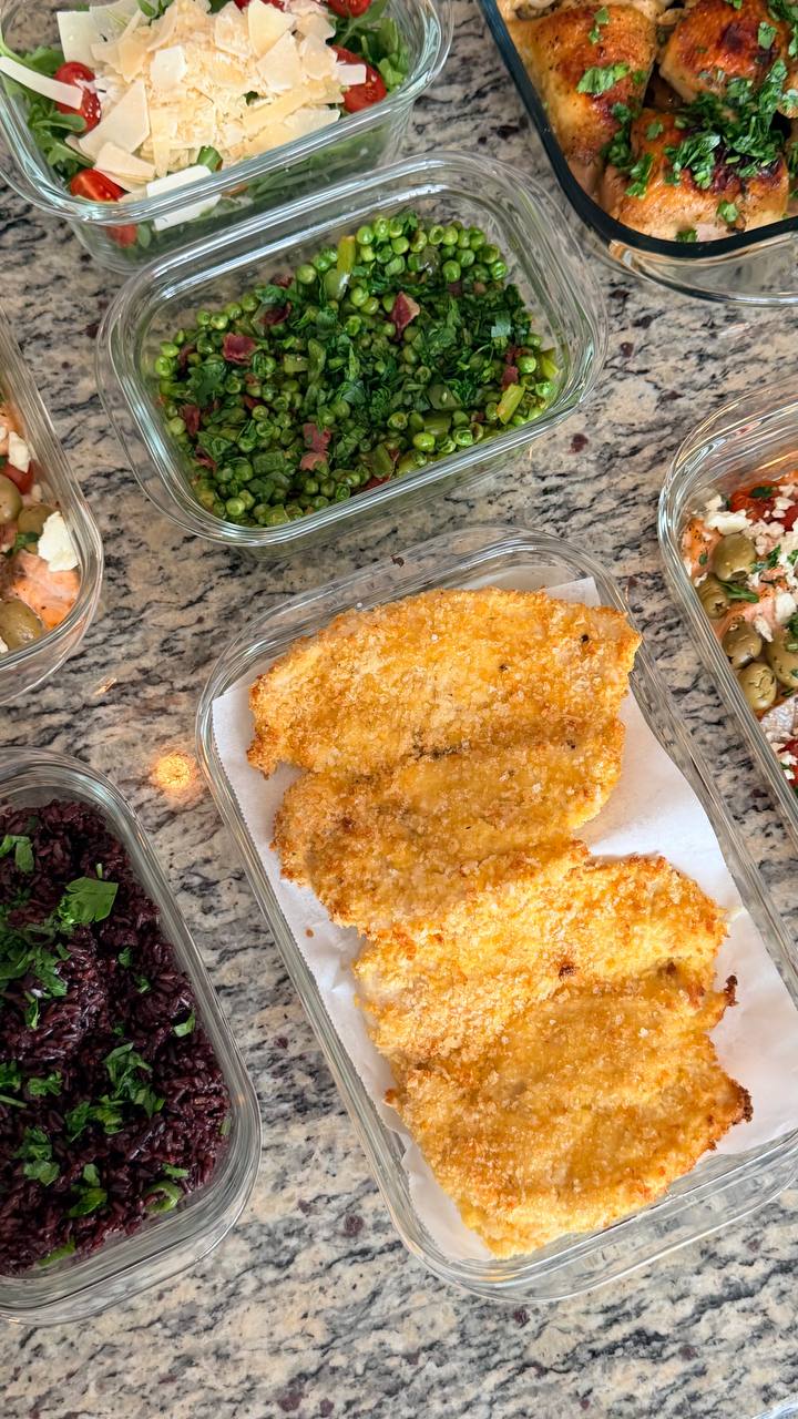 Glass meal prep containers filled with breaded chicken cutlets, black rice, seared bell peppers and peas, and a fresh arugula salad with parmesan.