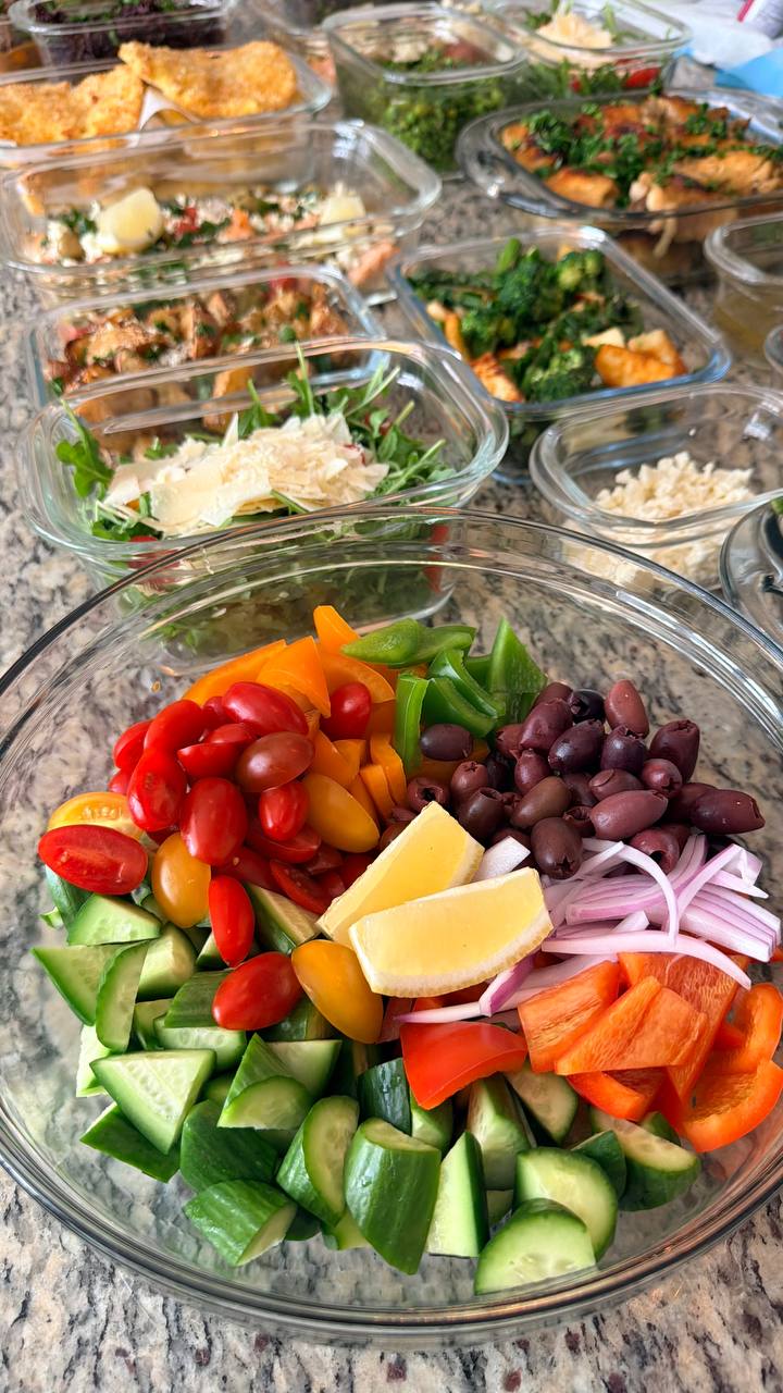 A large glass bowl filled with fresh Greek salad ingredients including cherry tomatoes, cucumber slices, kalamata olives, bell peppers, red onions, and lemon wedges.