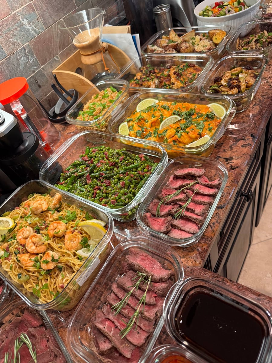 A variety of glass containers holding sliced filet mignon, curry coconut shrimp, seared asparagus, pancetta peas, and egg noodles on a granite countertop.
