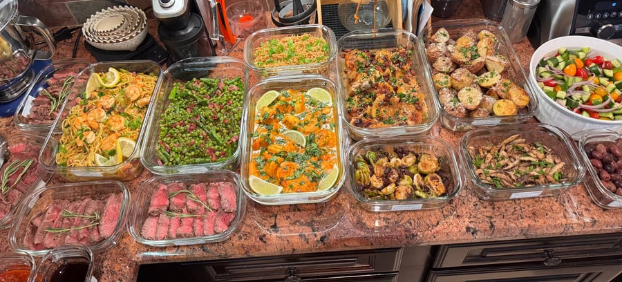 An overhead display of various gourmet dishes including filet mignon, shrimp noodles, roasted chicken, truffle potatoes, and Greek salad in glass containers.