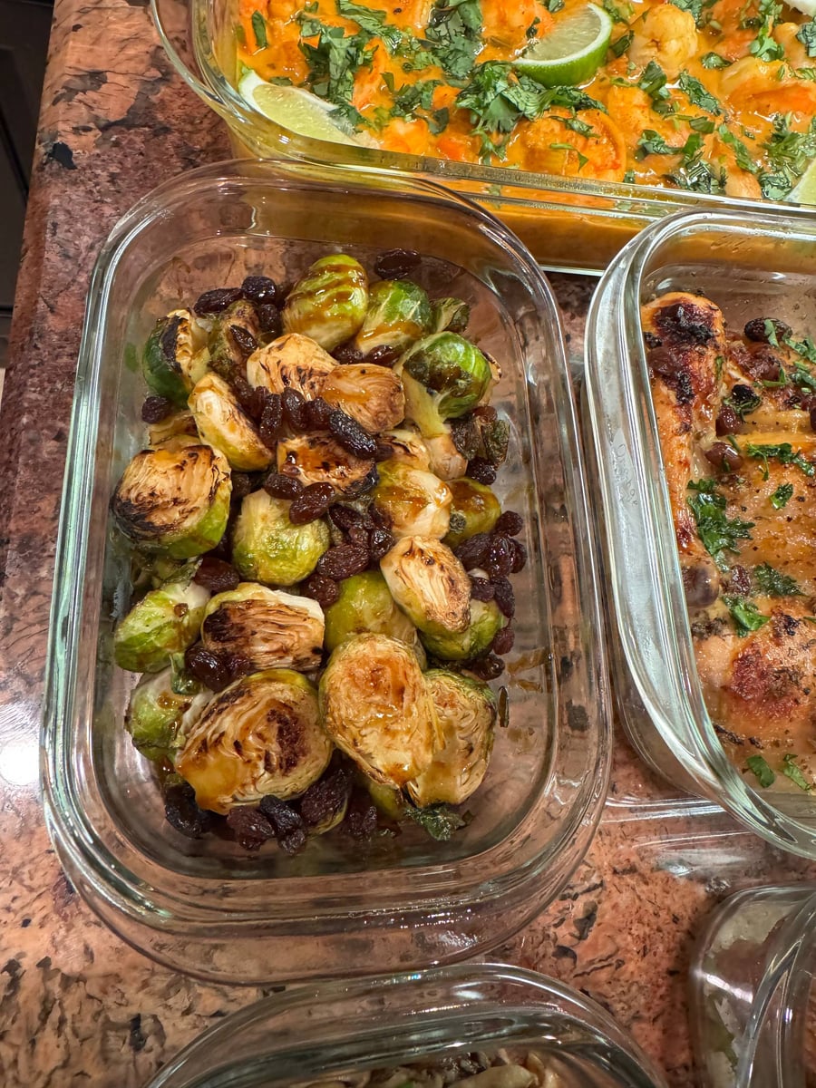 A glass container filled with roasted honey balsamic Brussel sprouts and dark raisins, sitting next to containers of chicken and curry coconut shrimp.