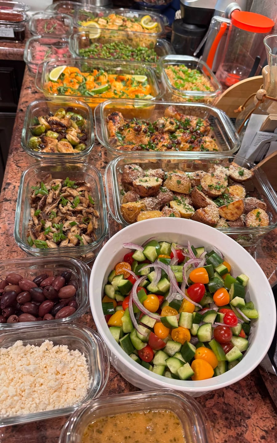 An overhead view of various glass containers featuring curry coconut shrimp, truffle parmesan baby potatoes, honey balsamic brussels sprouts, and a fresh Greek salad bowl.