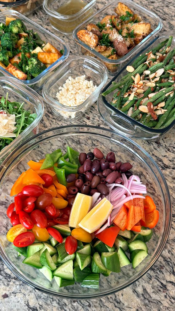 Glass containers holding classic Greek salad, green beans almondine, truffle parmesan baby potatoes, sauteed broccoli with paneer, and a bowl of fresh feta cheese.