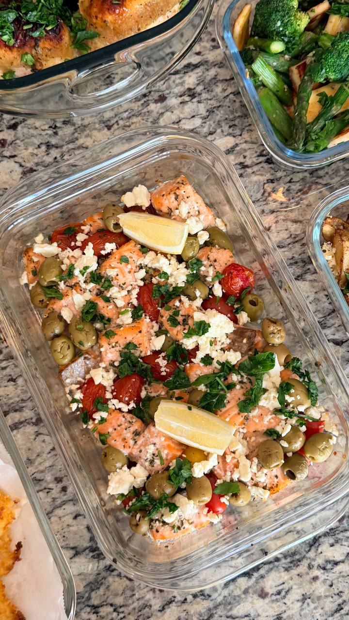 A glass container holds Mediterranean baked salmon bites topped with crumbled feta cheese, green olives, cherry tomatoes, fresh herbs, and lemon wedges.