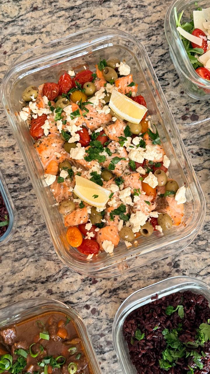 Glass container filled with Mediterranean baked salmon bites topped with green olives, crumbled feta cheese, fresh herbs, and bright lemon wedges for garnish.