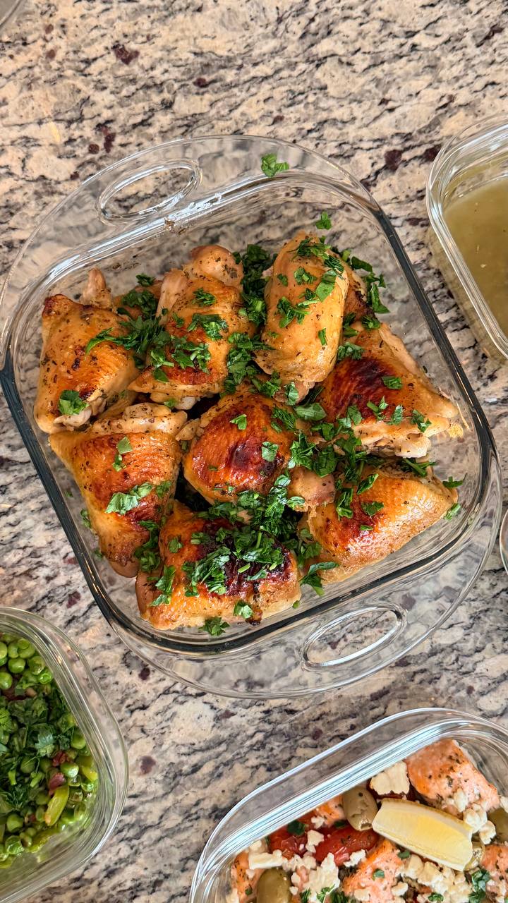 A glass baking dish containing golden-brown roasted chicken thighs garnished with fresh green herbs, displayed on a granite countertop alongside other prepared sides.