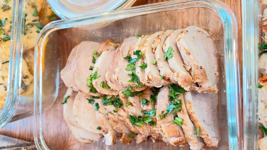 A glass storage container filled with neatly arranged slices of paprika-baked pork tenderloin garnished with a generous sprinkling of fresh chopped green herbs.
