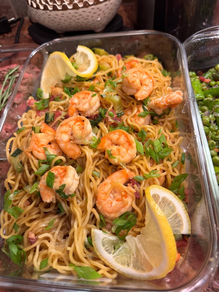 A glass container filled with shrimp and bok choy egg noodles, garnished with fresh green scallions and bright lemon wedges for a vibrant presentation.