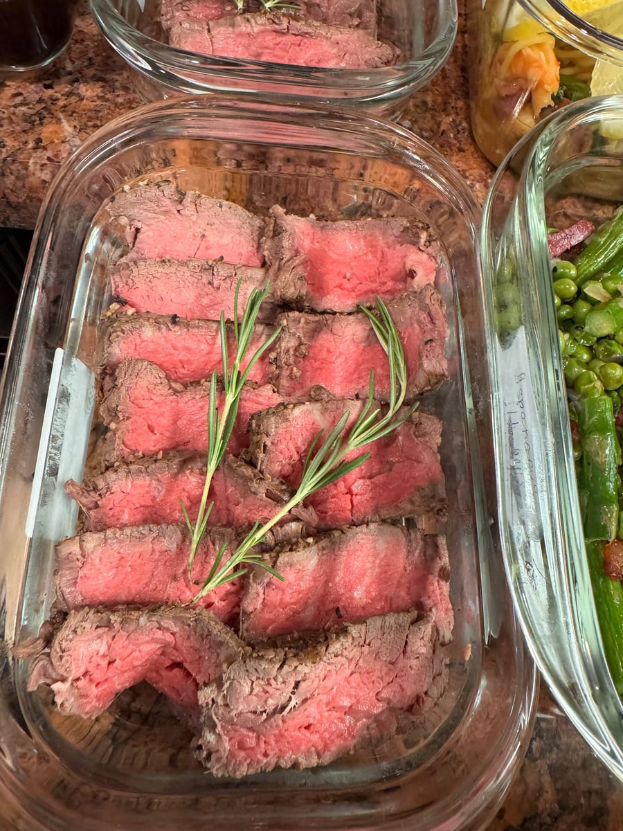A glass container features perfectly sliced medium-rare filet mignon steaks garnished with fresh rosemary sprigs, arranged neatly for a gourmet meal preparation.