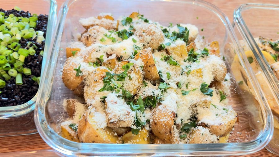 A glass container holds golden baked baby potatoes dusted with fluffy parmesan cheese and fresh green herbs, displayed alongside black rice with scallions.