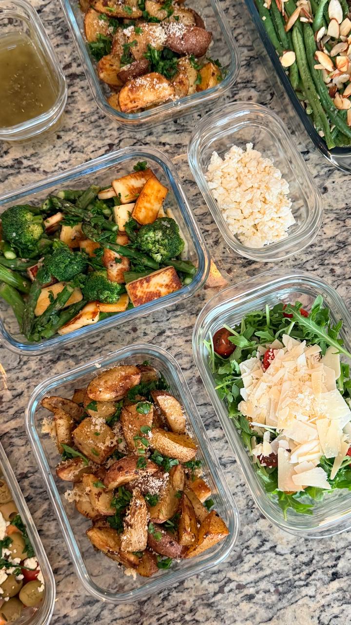 Glass containers featuring truffle parmesan baby potatoes, sautéed broccoli with paneer, and an arugula salad topped with shaved parmesan and fresh tomatoes.