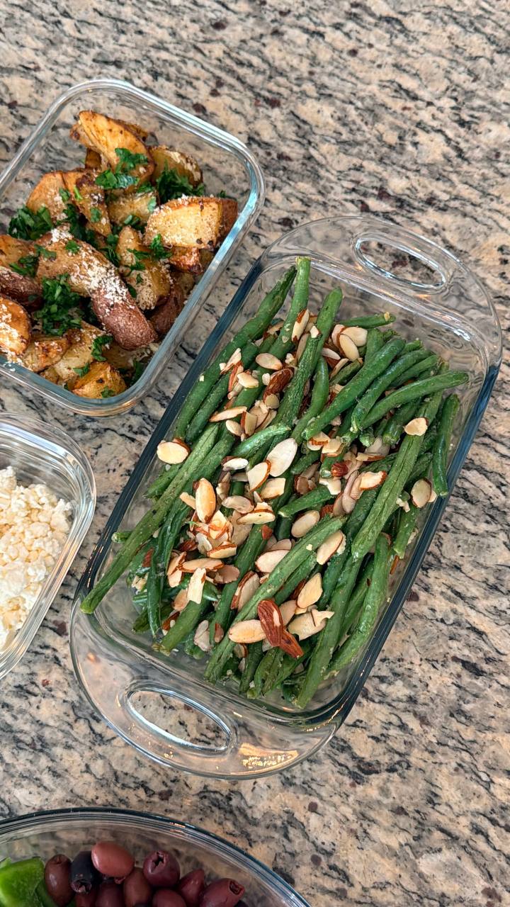 Glass containers holding roasted truffle parmesan baby potatoes and fresh green beans almondine with toasted almond slivers, arranged nicely on a granite countertop.